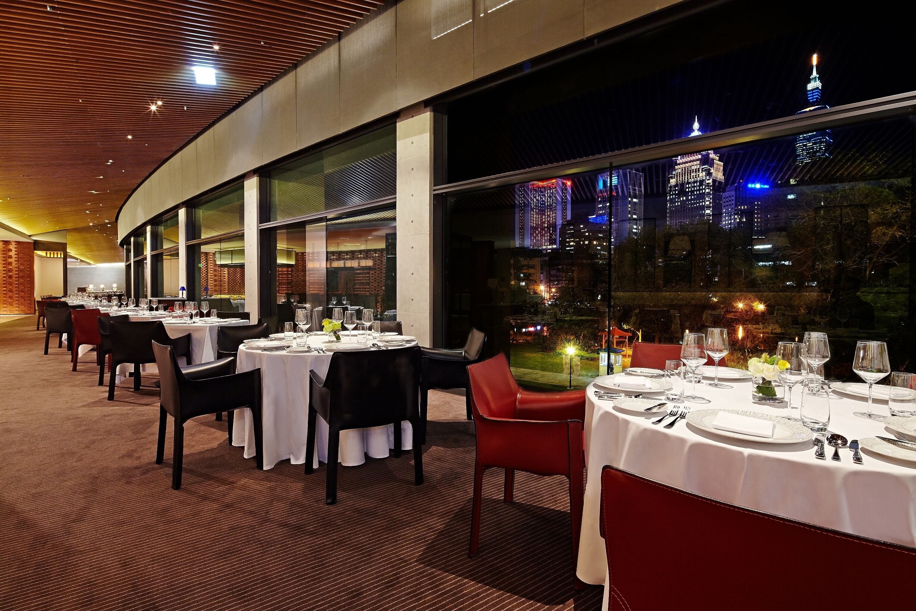 A restaurant with multiple tables set for dining, large windows offering a view of the cityscape at night.