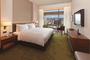 Habitación de hotel con una cama grande, un escritorio, una silla y vistas de la ciudad a través de un amplio ventanal.