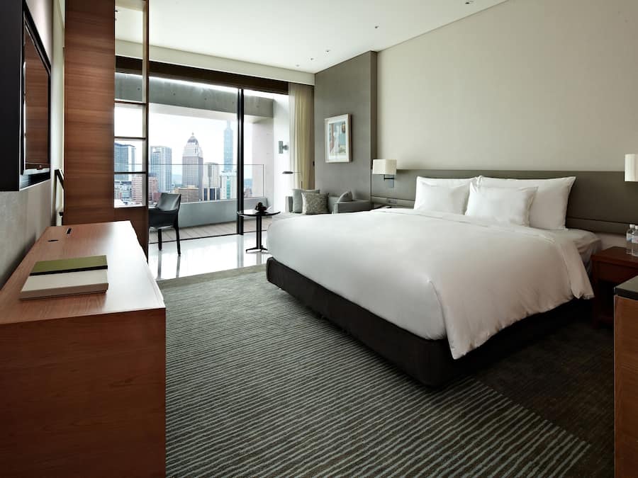 A modern hotel room with a large bed, a desk, a chair, and a view of the city skyline.