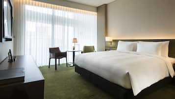 A hotel room with a large bed, a desk, a chair, a small table with a lamp, and a large window with sheer curtains.