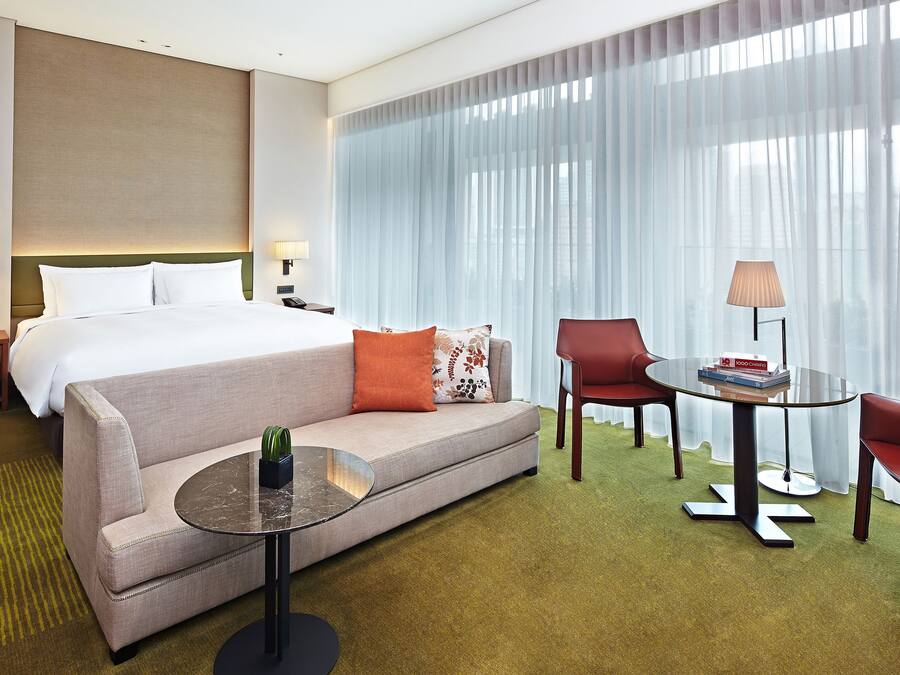 A hotel room with a bed, a sofa, a coffee table, two chairs, a lamp, and a nightstand with a clock and a phone.