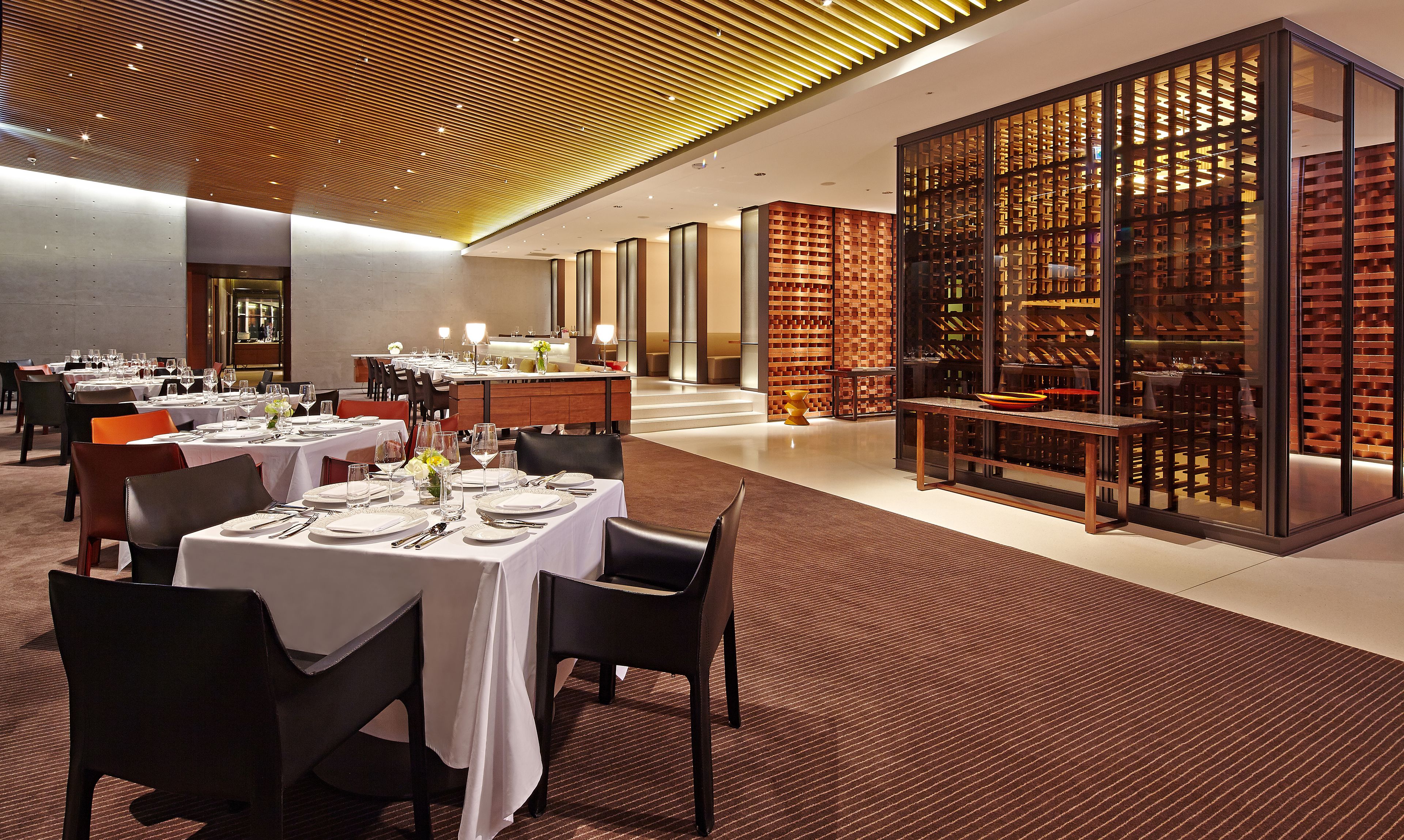 A dining area with tables set for service, a wine cellar visible through a glass partition, and a modern ceiling design.