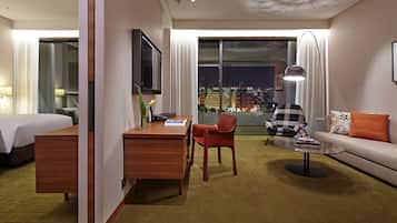 A hotel room with a bed, a desk, a chair, a sofa, and a view of the city at night.