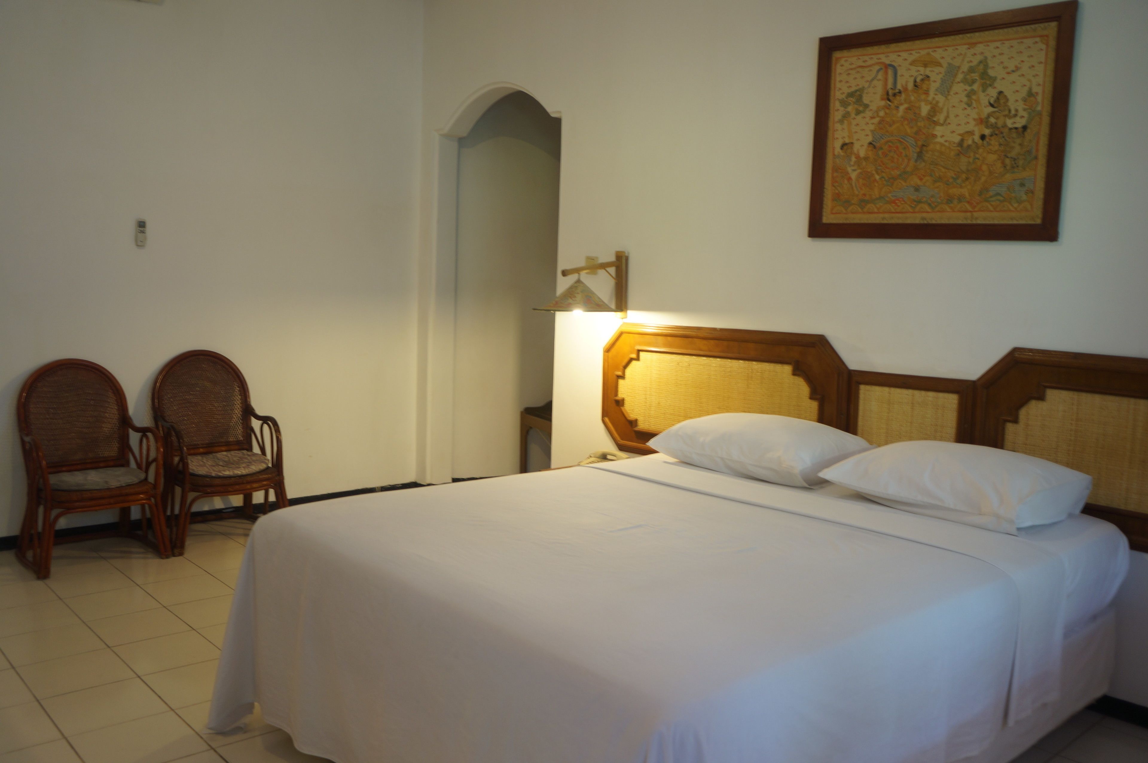 A hotel room with a large bed, two wooden chairs, a wall-mounted lamp, and a framed picture.
