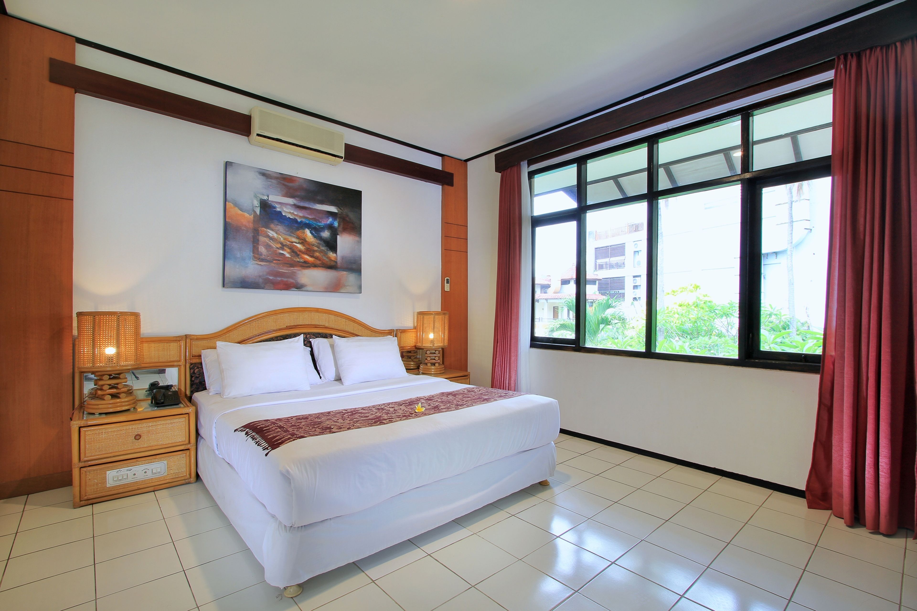 A hotel room with a large bed, a nightstand with a lamp, a painting on the wall, and a window with curtains.