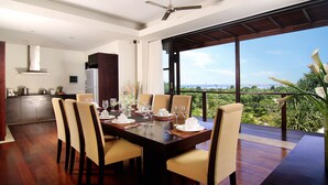 A dining area with a large table set for six, a kitchen in the background, and a balcony with a view of the outdoors. - Temple Hill Residence Villa (Jimbaran)