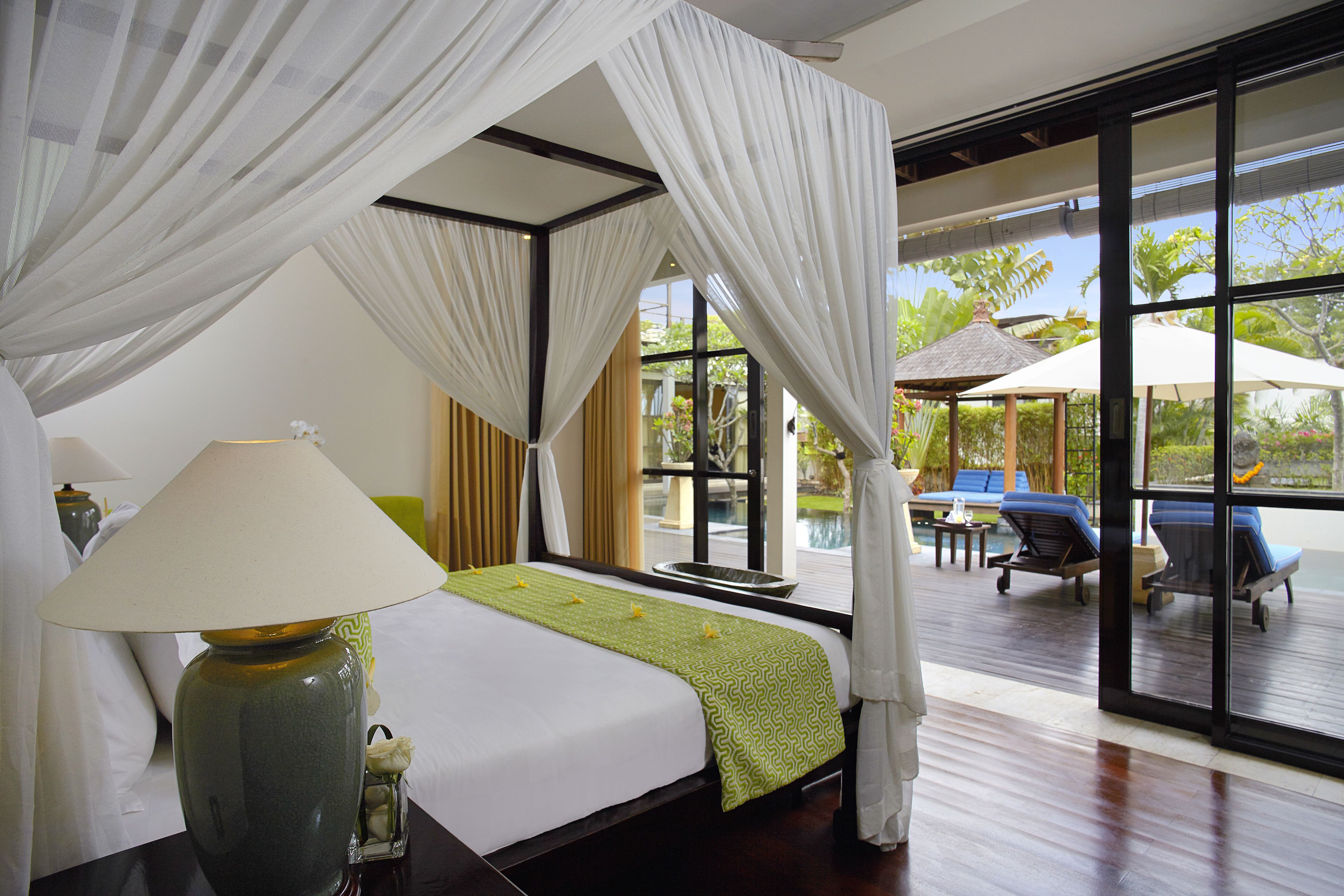 A hotel room with a large bed, a nightstand, a lamp, and a view of the outdoor pool area.