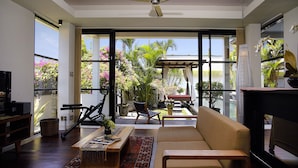 A spacious living room with a sofa, coffee table, and a ceiling fan. Large windows offer a view of a garden with plants and a pergola. - Temple Hill Residence Villa (Jimbaran)