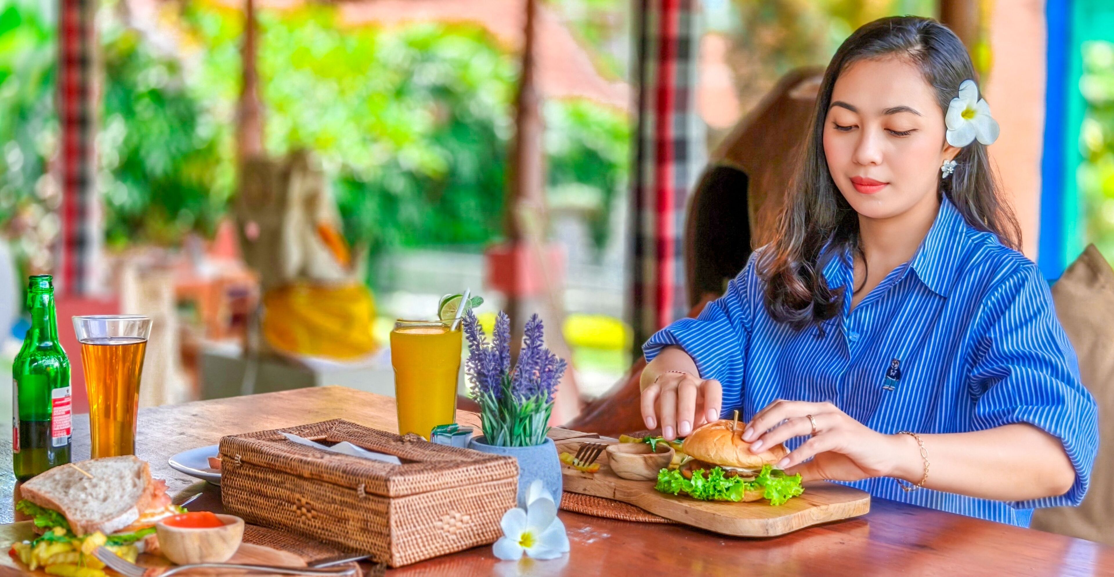 A person assembling a sandwich at a wooden table with a drink and a bottle of beer.