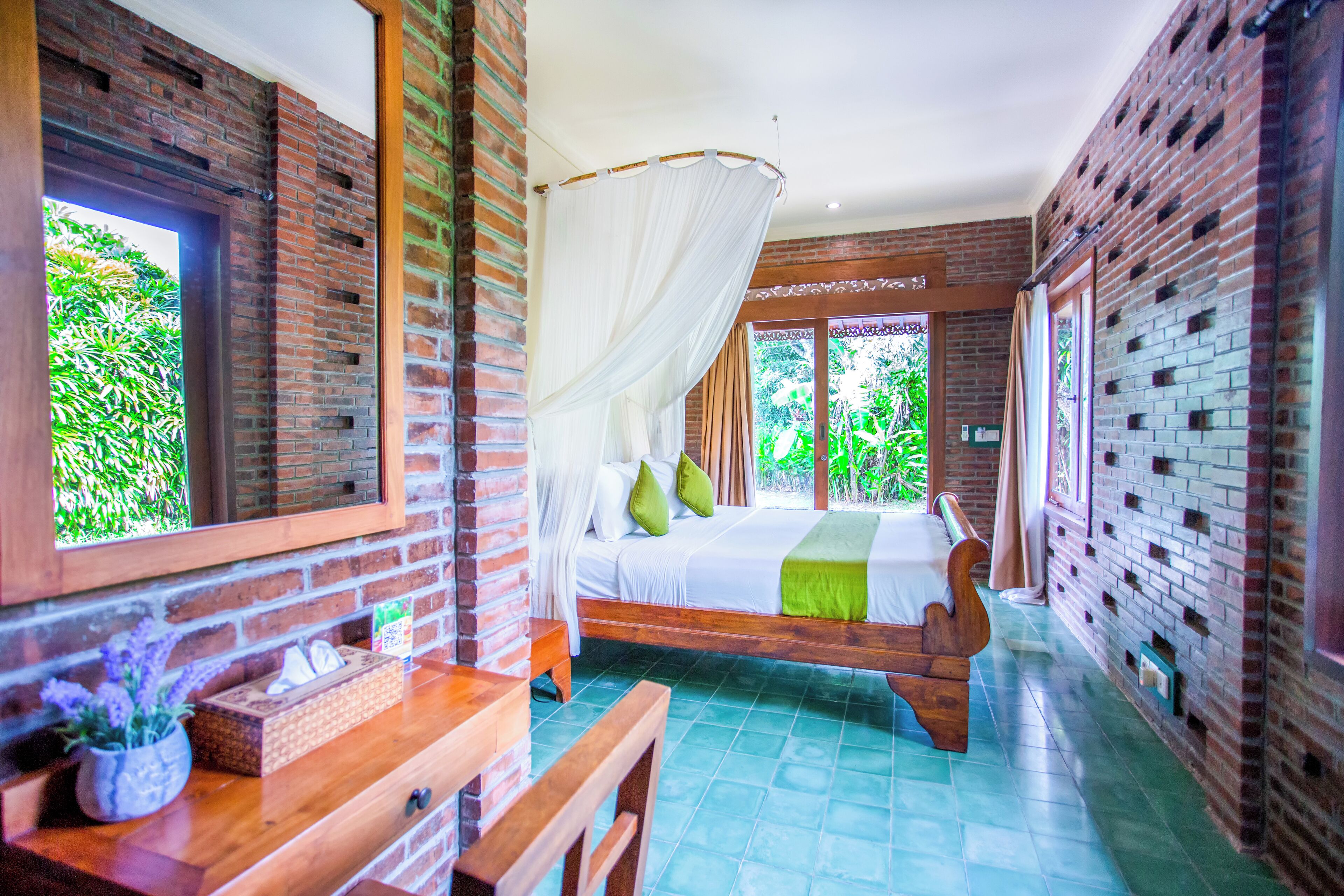 Un dormitorio con cama, mesita de noche, banqueta y ventana con vista a follaje.