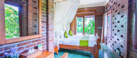 Un dormitorio con cama, mesita de noche, banqueta y ventana con vista a follaje.