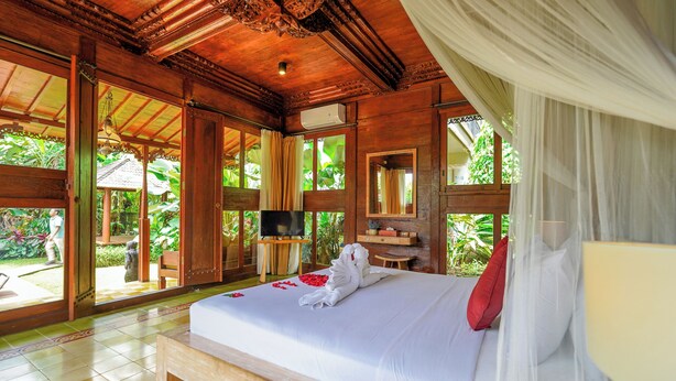 A large bed with a white bedspread, a red pillow, and a towel. There is a television on a wooden stand, a small table, and a chair. The room has wooden walls and a wooden ceiling. - Ubud Heaven Penestanan (Ubud)