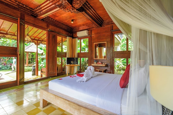 A large bed with a white bedspread, a red pillow, and a towel. There is a television on a wooden stand, a small table, and a chair. The room has wooden walls and a wooden ceiling. - Ubud Heaven Penestanan (Ubud)