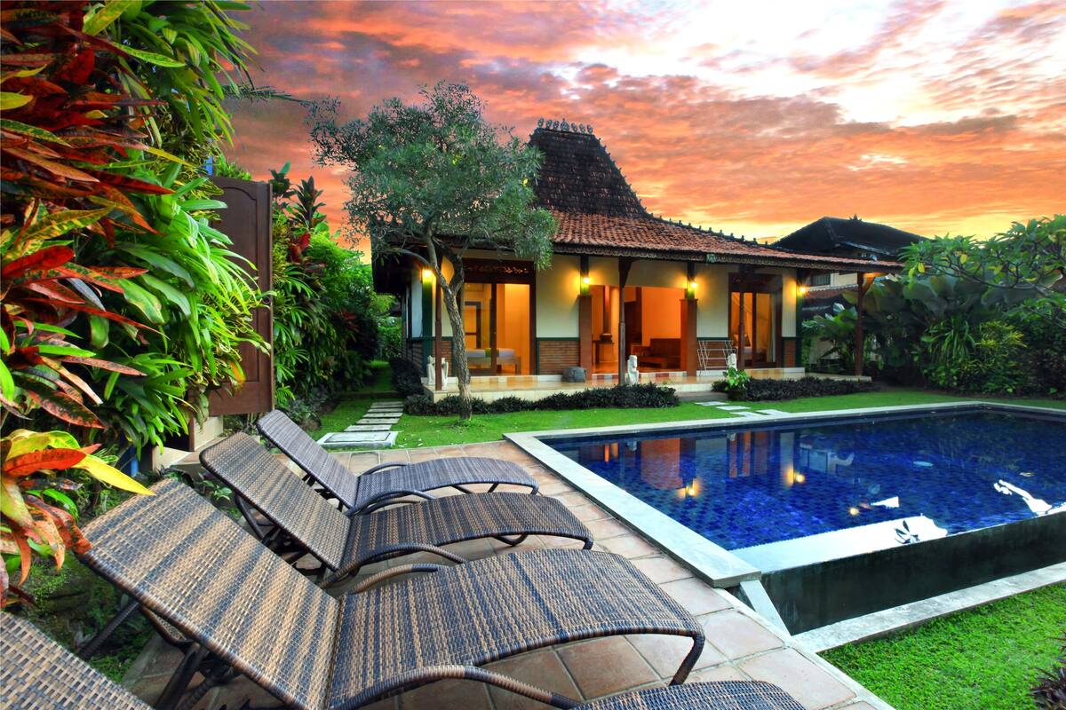 A house with a swimming pool, surrounded by lush greenery and a sunset sky.