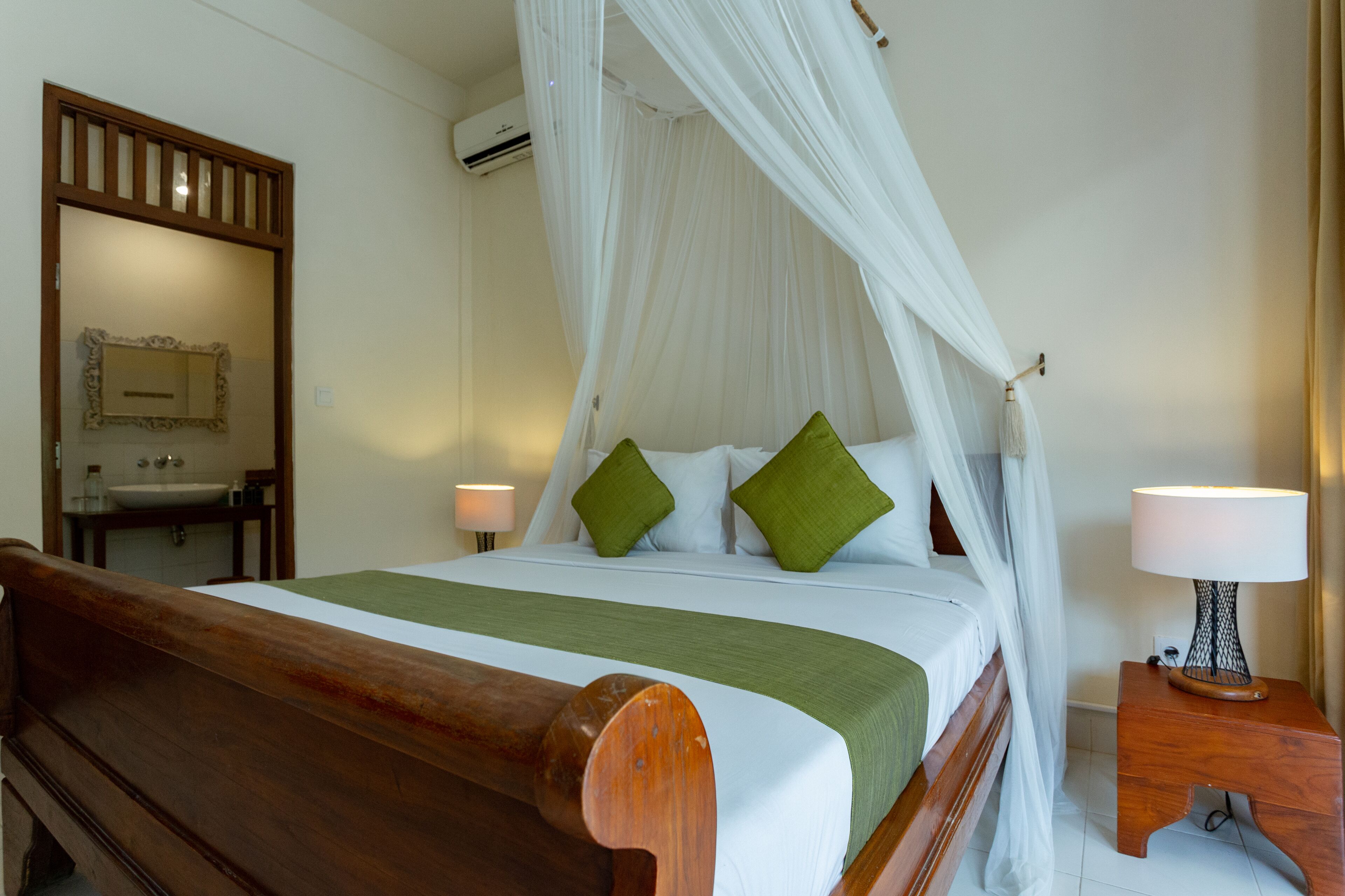 Una habitación de hotel con cama de madera, dosel blanco, almohadas verdes, mesita de noche con lámpara y un espejo en la pared.