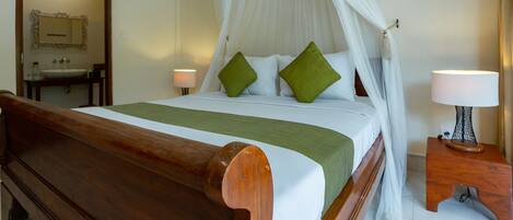 A hotel room with a wooden bed, white canopy bed, green pillows, bedside table with a lamp, and a mirror on the wall.