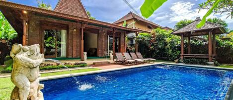 A swimming pool with a stone statue, a covered patio, and a traditional house with a tiled roof.