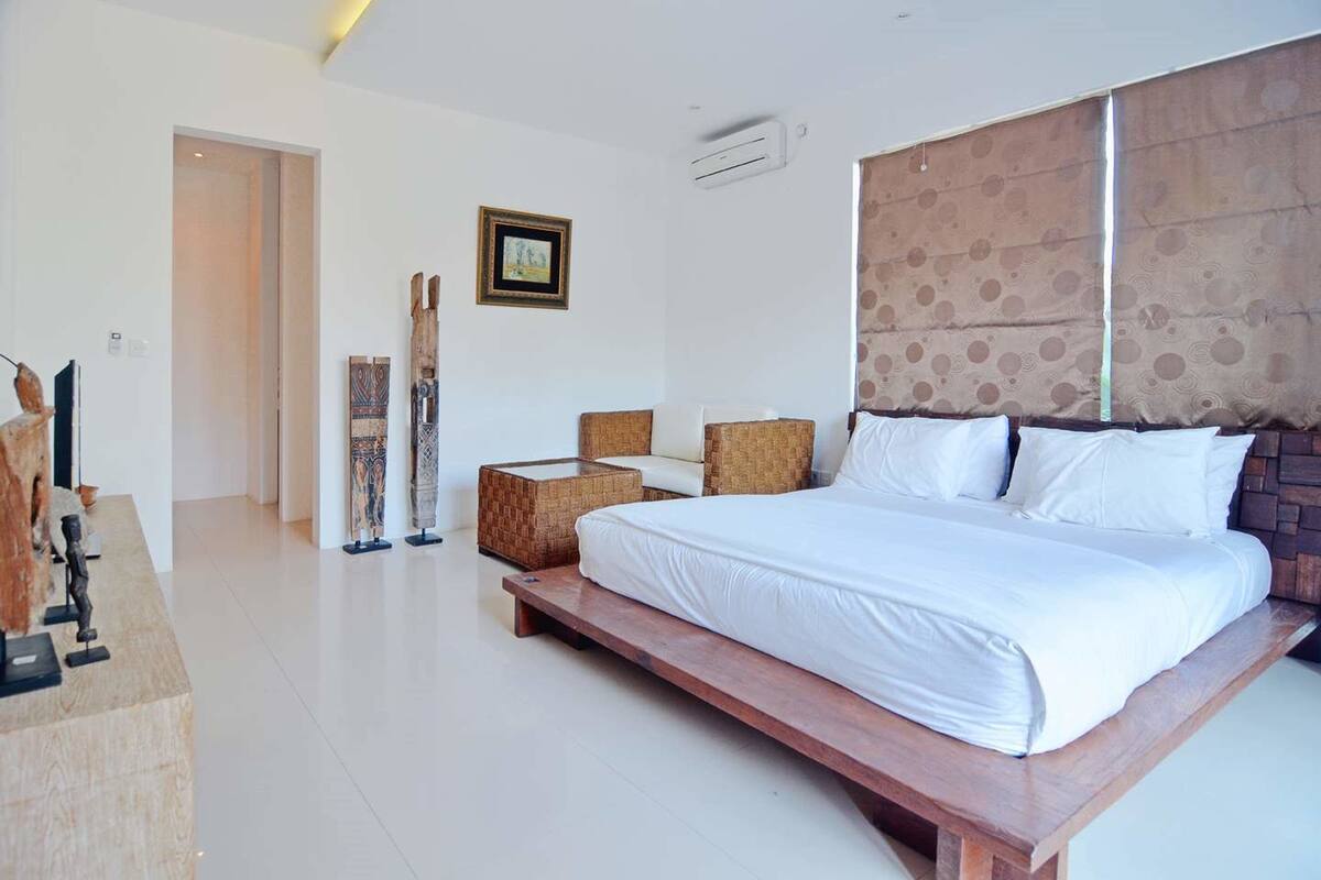 A modern bedroom with a large bed, a wooden nightstand, a small table, and a chair. There is a framed picture on the wall and an air conditioning unit above the bed.