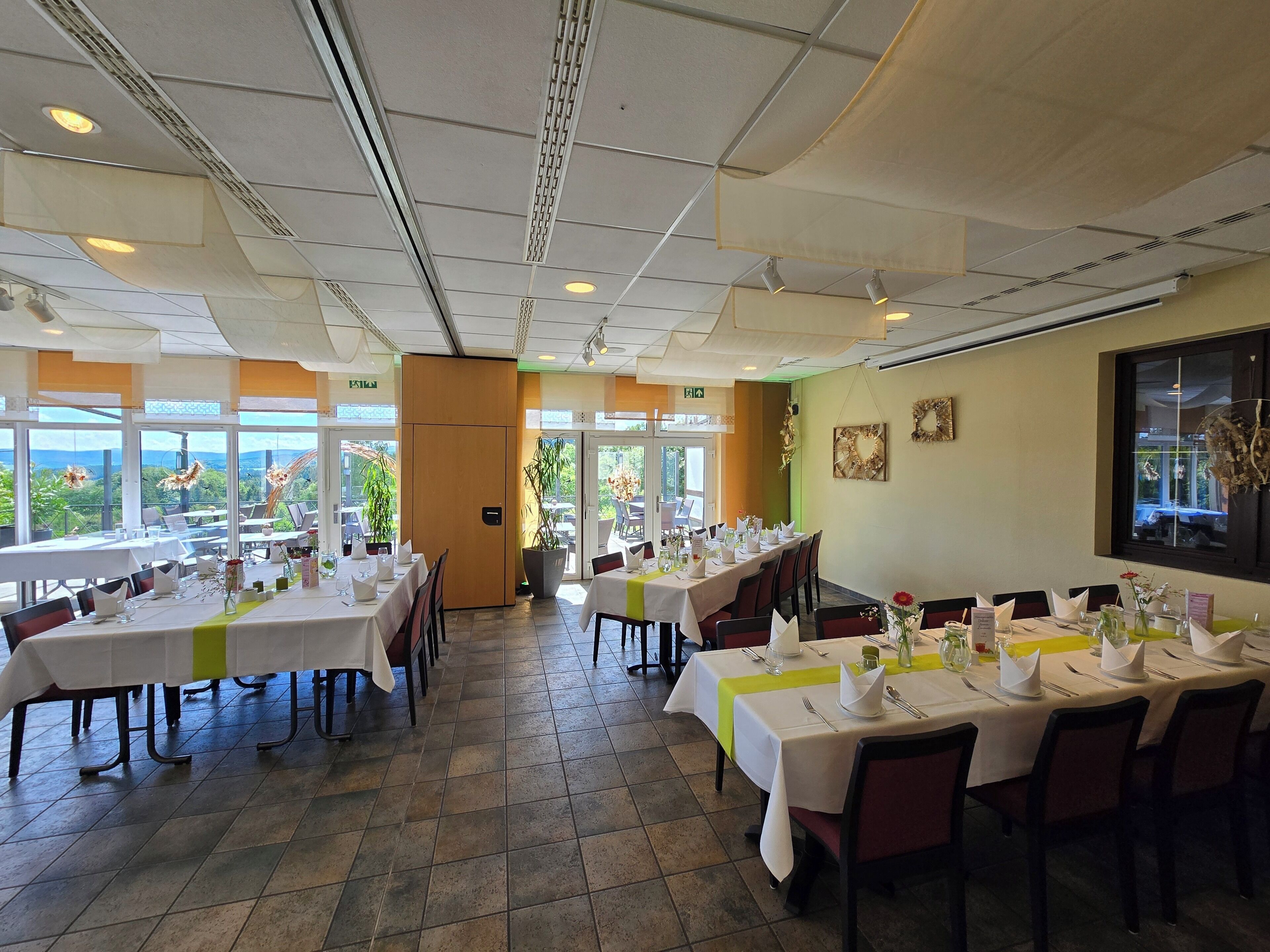 A restaurant with multiple tables set for dining, large windows offering a view, and a high ceiling with recessed lighting.