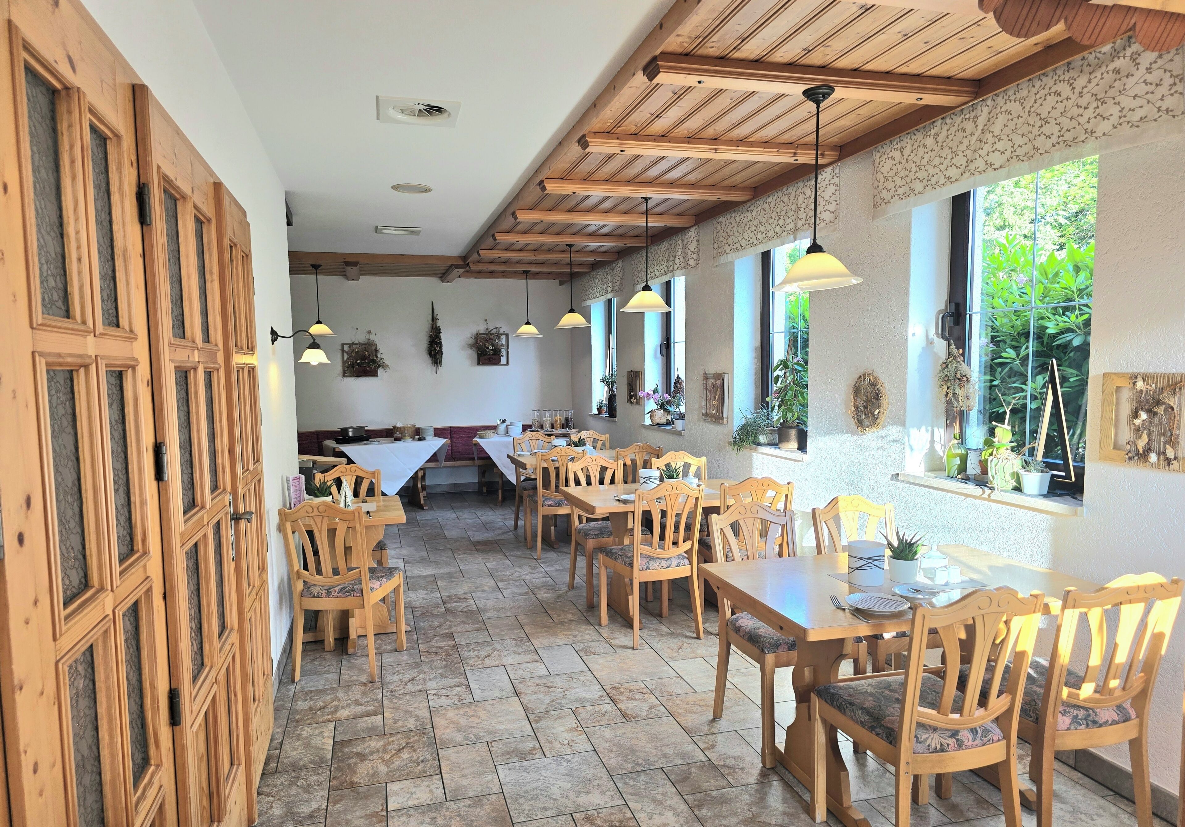 Un comedor con mesas y sillas de madera, una cocina al fondo y ventanas grandes con plantas.