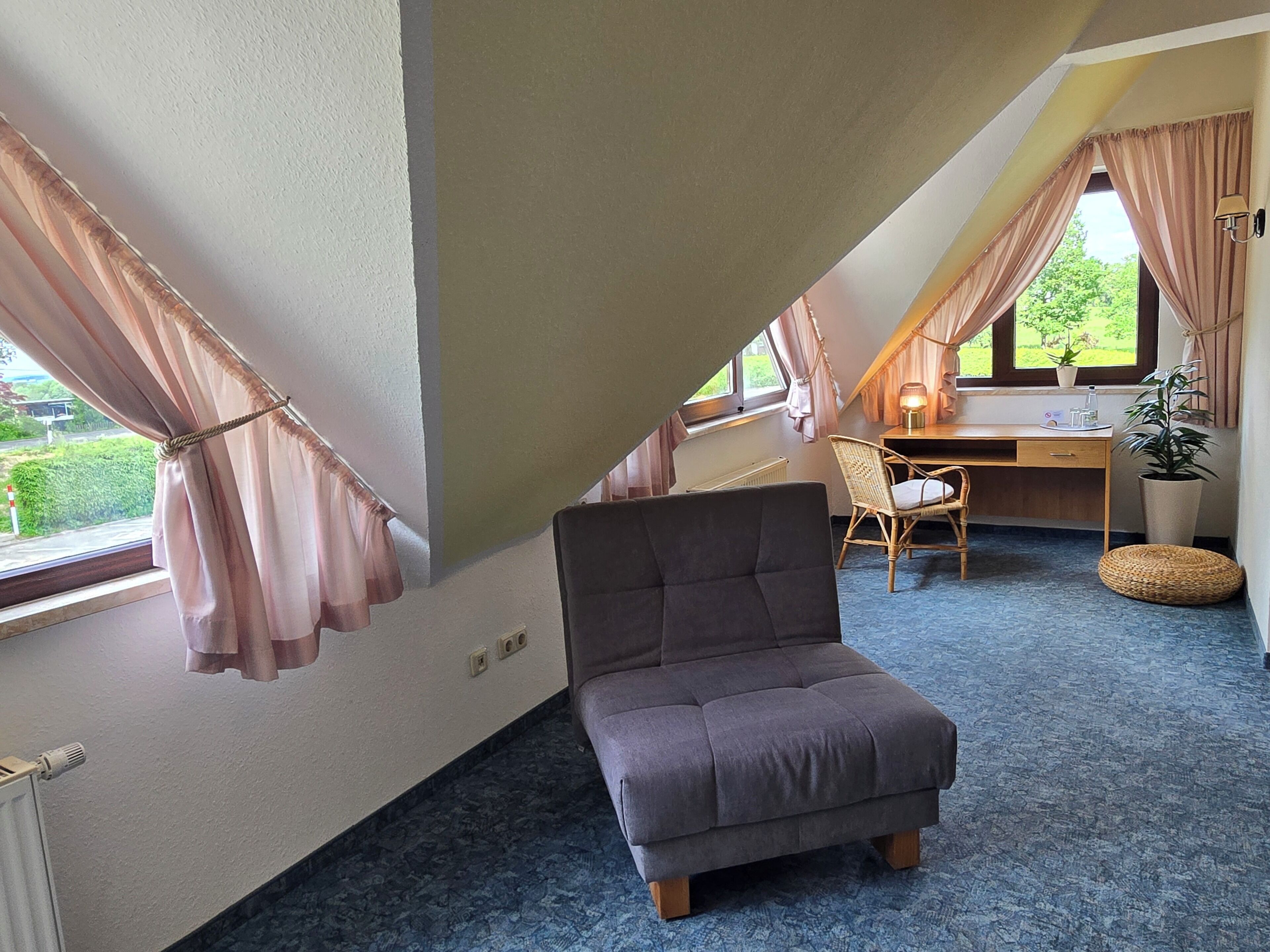 Una habitación con techo inclinado, una cama individual, un escritorio con silla, una planta en maceta y una ventana con cortinas.