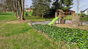 Children can play on the playground's slide and swings in a lush, grassy area surrounded by trees and residential buildings.