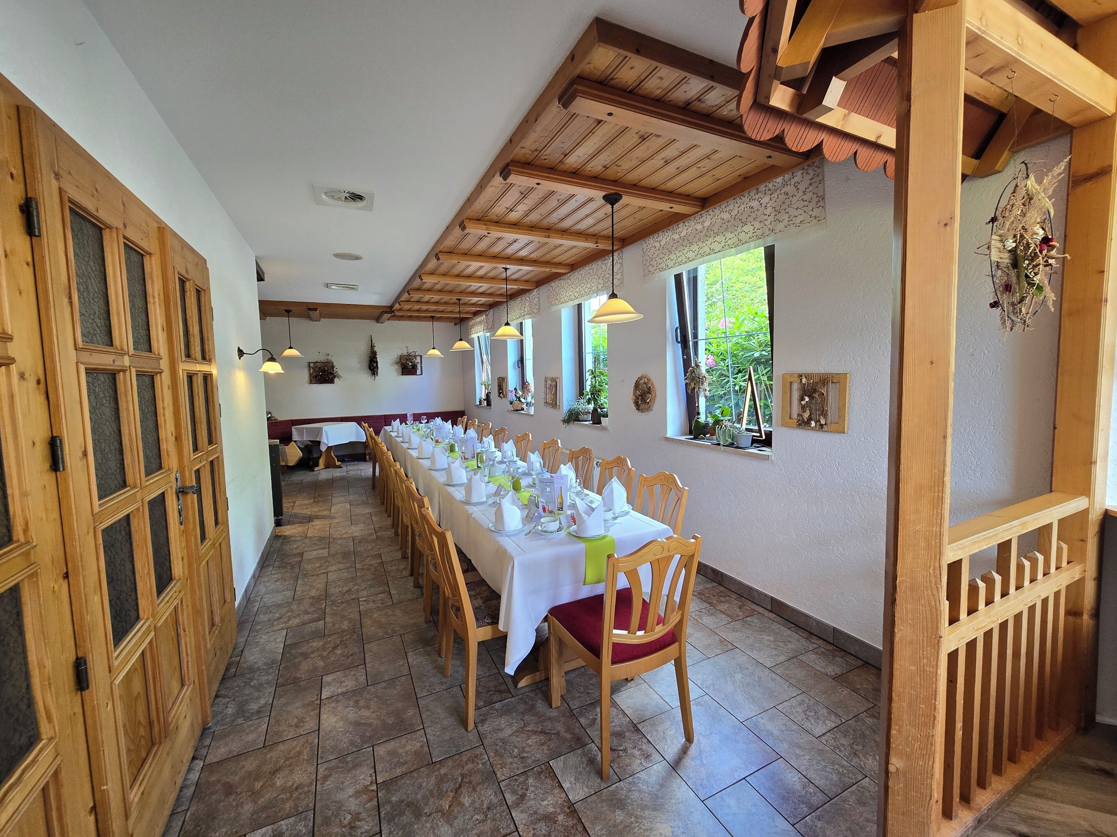 Una larga mesa de comedor con sillas, vigas de madera en el techo y ventanas con plantas.