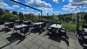 Patio esterno con tavoli e sedie, vista su spazi verdi e colline, e cielo azzurro con nuvole.
