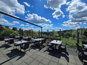 Un patio exterior con mesas y sillas, vistas a zonas verdes y colinas, y un cielo azul despejado con nubes.