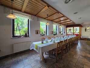 Una larga mesa de comedor con sillas, vigas de madera en el techo y ventanas con plantas.