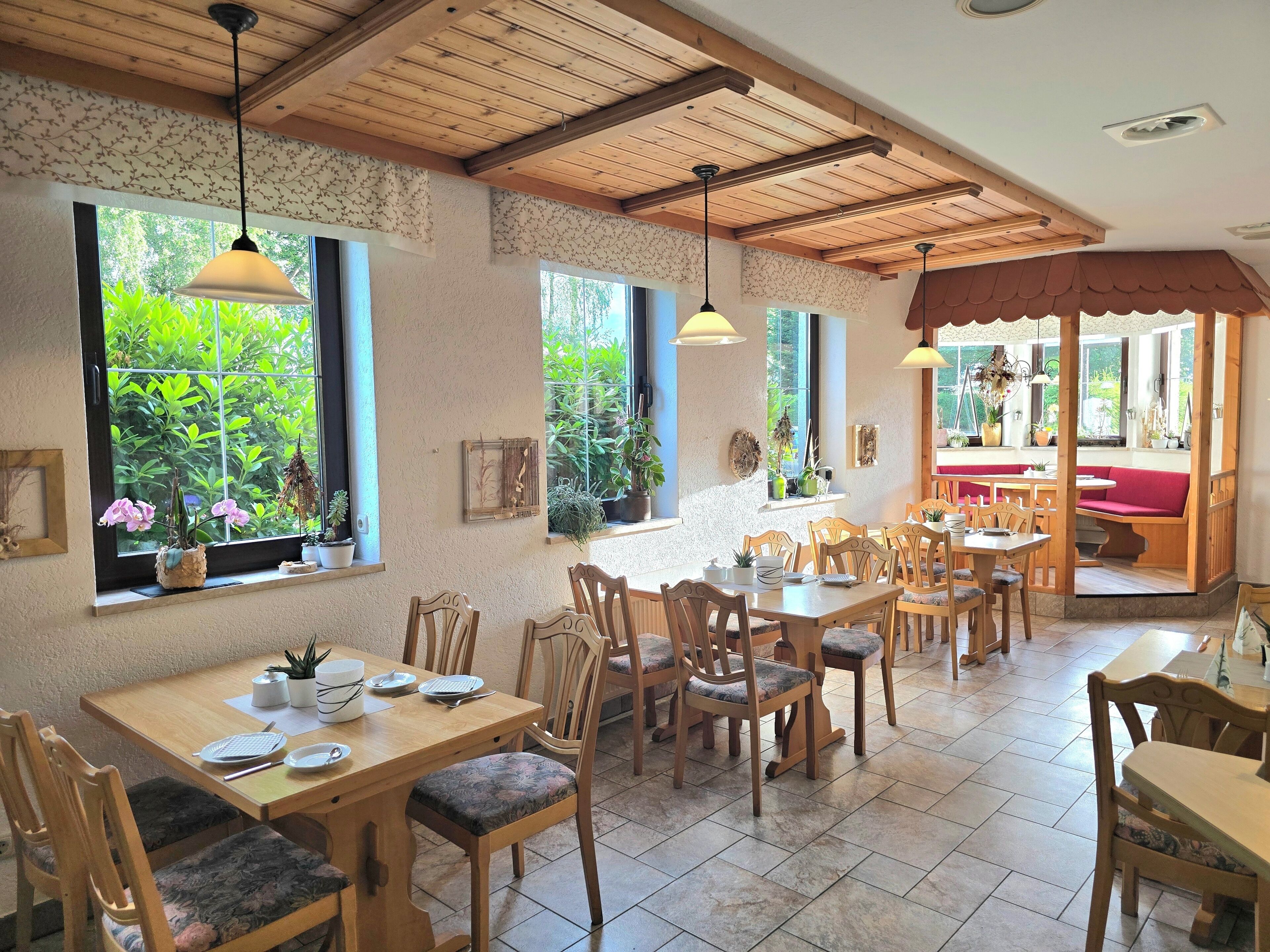 Un comedor con mesas y sillas de madera, ventanas amplias con plantas y techo de madera.