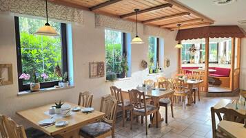 Un comedor con mesas y sillas de madera, ventanas amplias con plantas y techo de madera.