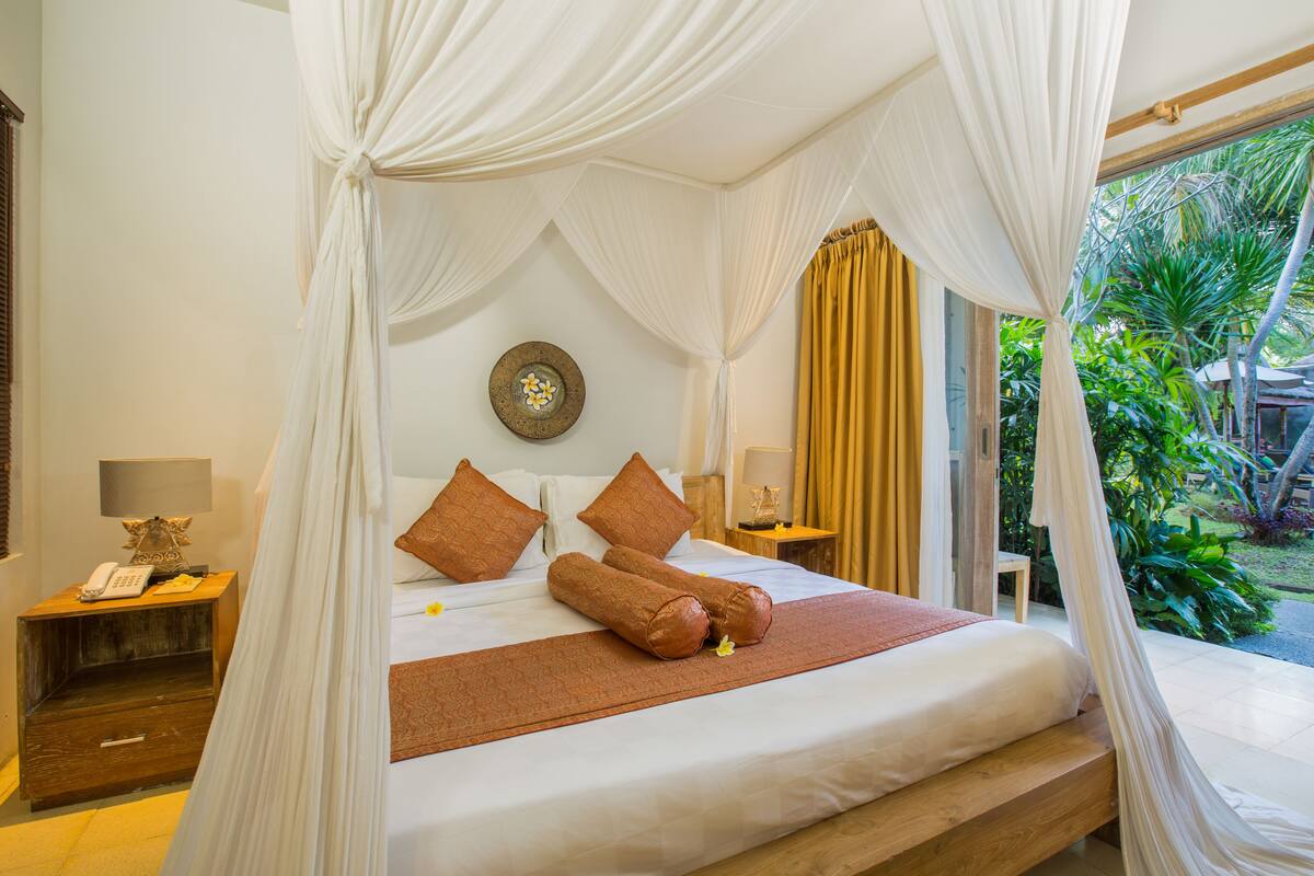 A room with a bed, a nightstand, a lamp, and a view of greenery outside.