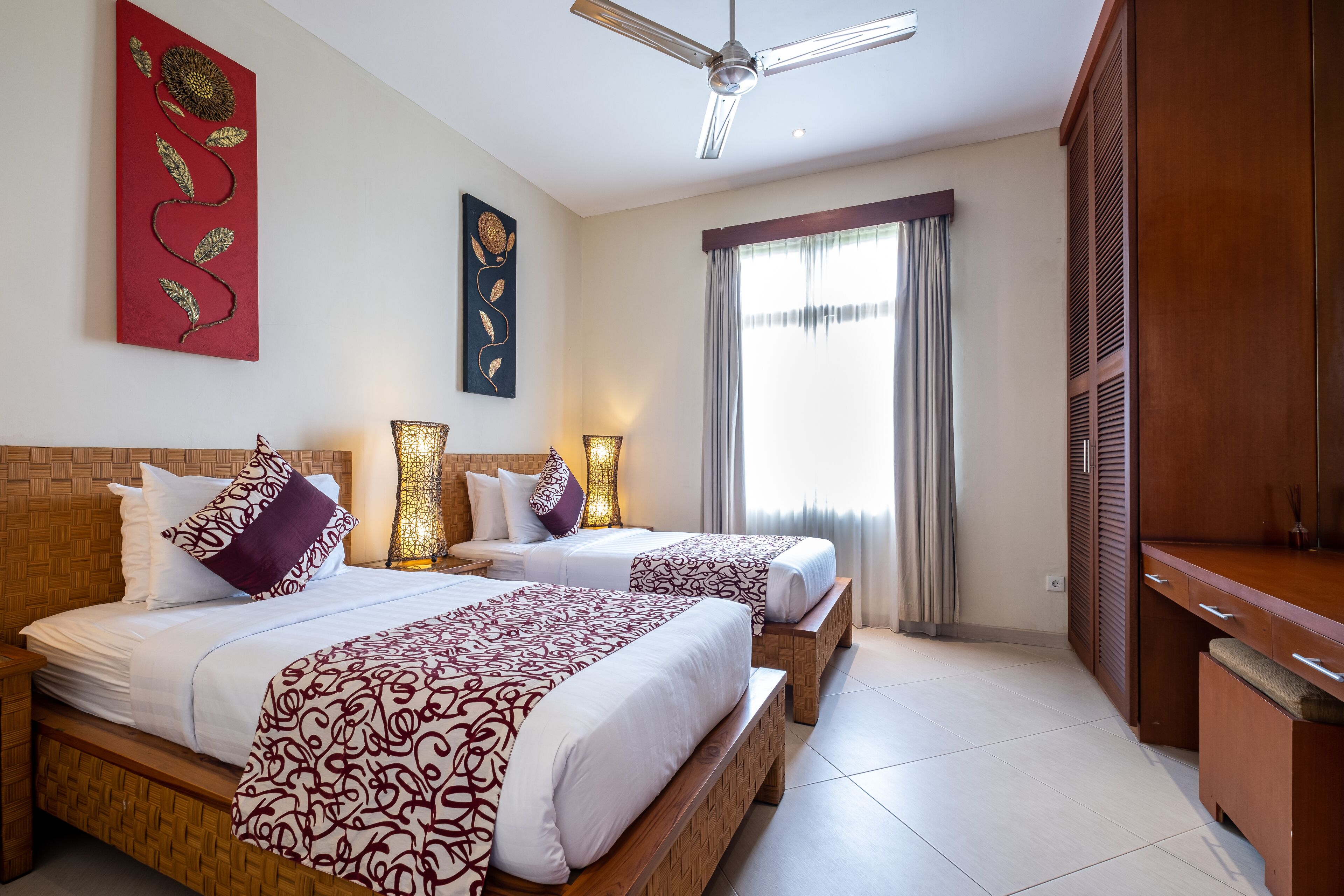 Habitación de hotel con dos camas, ventilador de techo, ventana grande con cortinas y armario de madera.