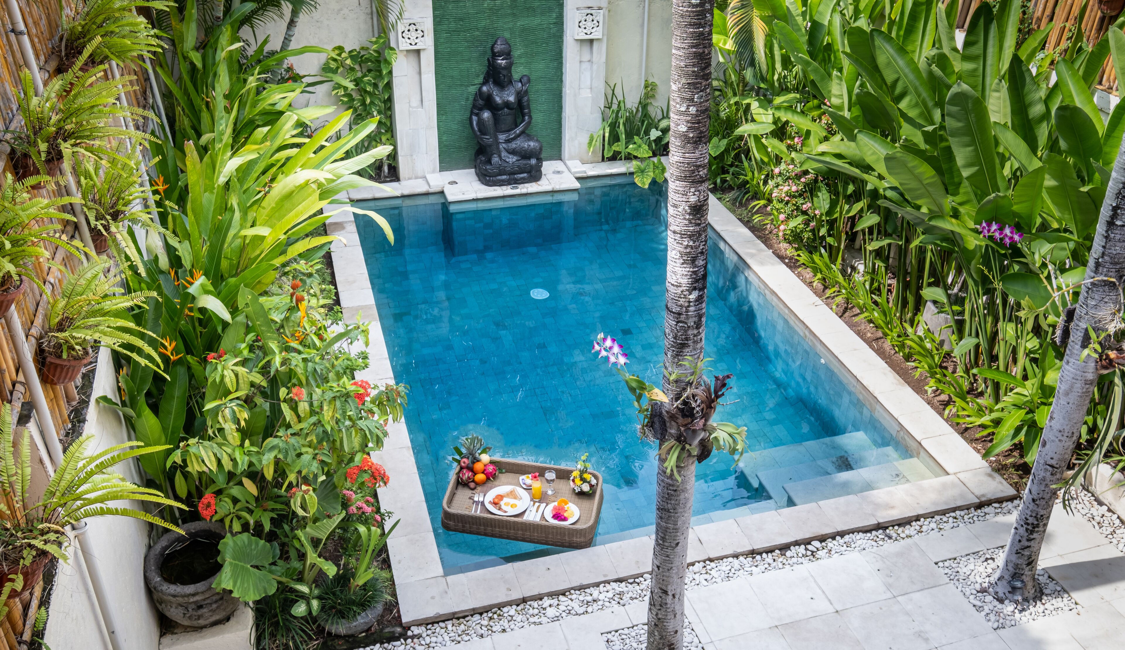 Ein Pool, umgeben von üppiger Vegetation, einer Statue und einem Frühstücksbuffet.