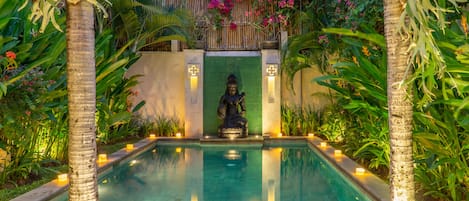 A serene pool area with a statue, surrounded by lush greenery and palm trees, with candles lining the pool's edge.