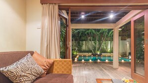 A room with a brown sofa, a wooden table, and a view of an outdoor pool area with plants and candles. - Villa Seriska Seminyak Bali (Seminyak)