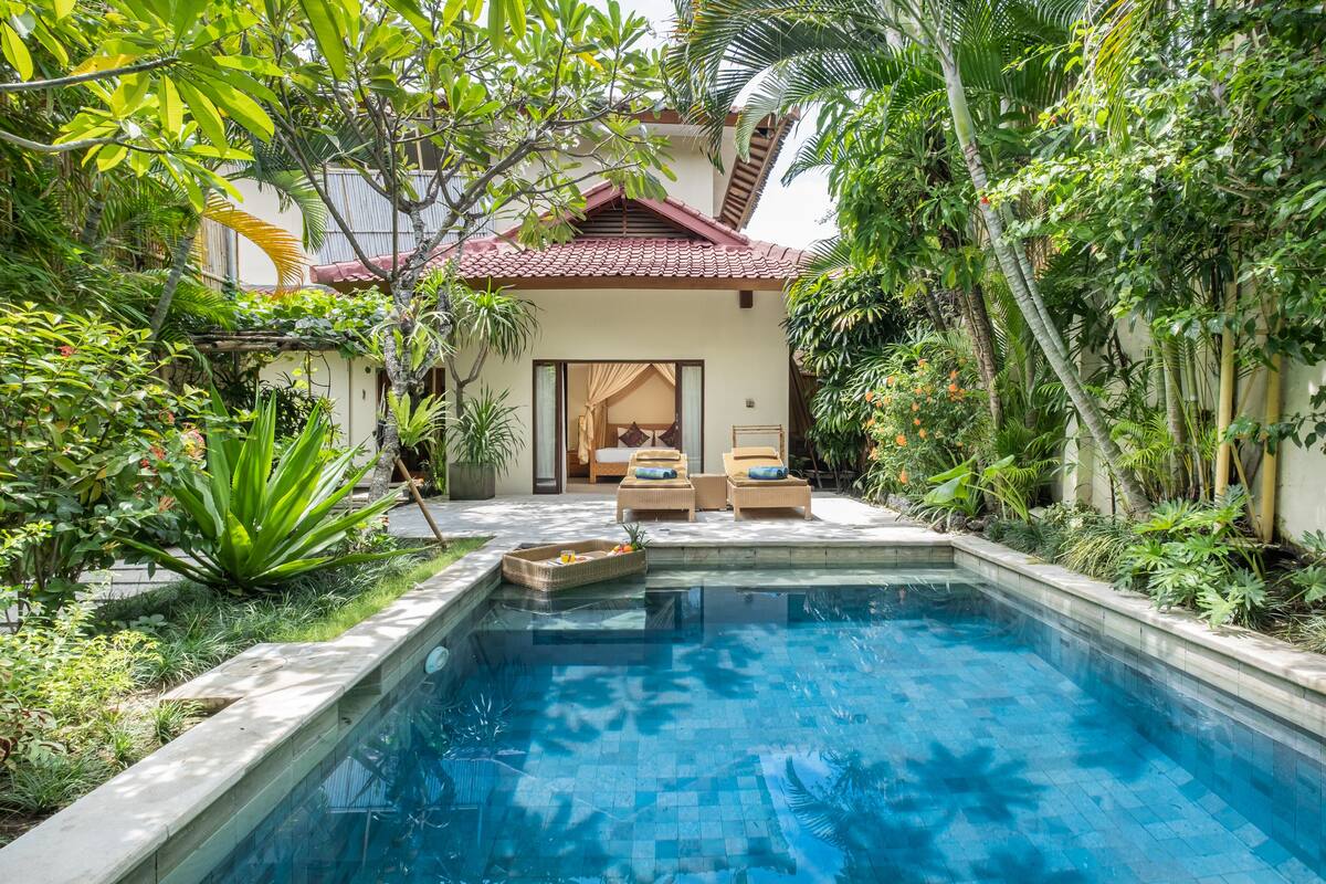 Un espace piscine agrémenté d’un lit et de chaises longues, entouré d’une végétation luxuriante et bordé d’un bâtiment dont le toit est recouvert de tuiles rouges.