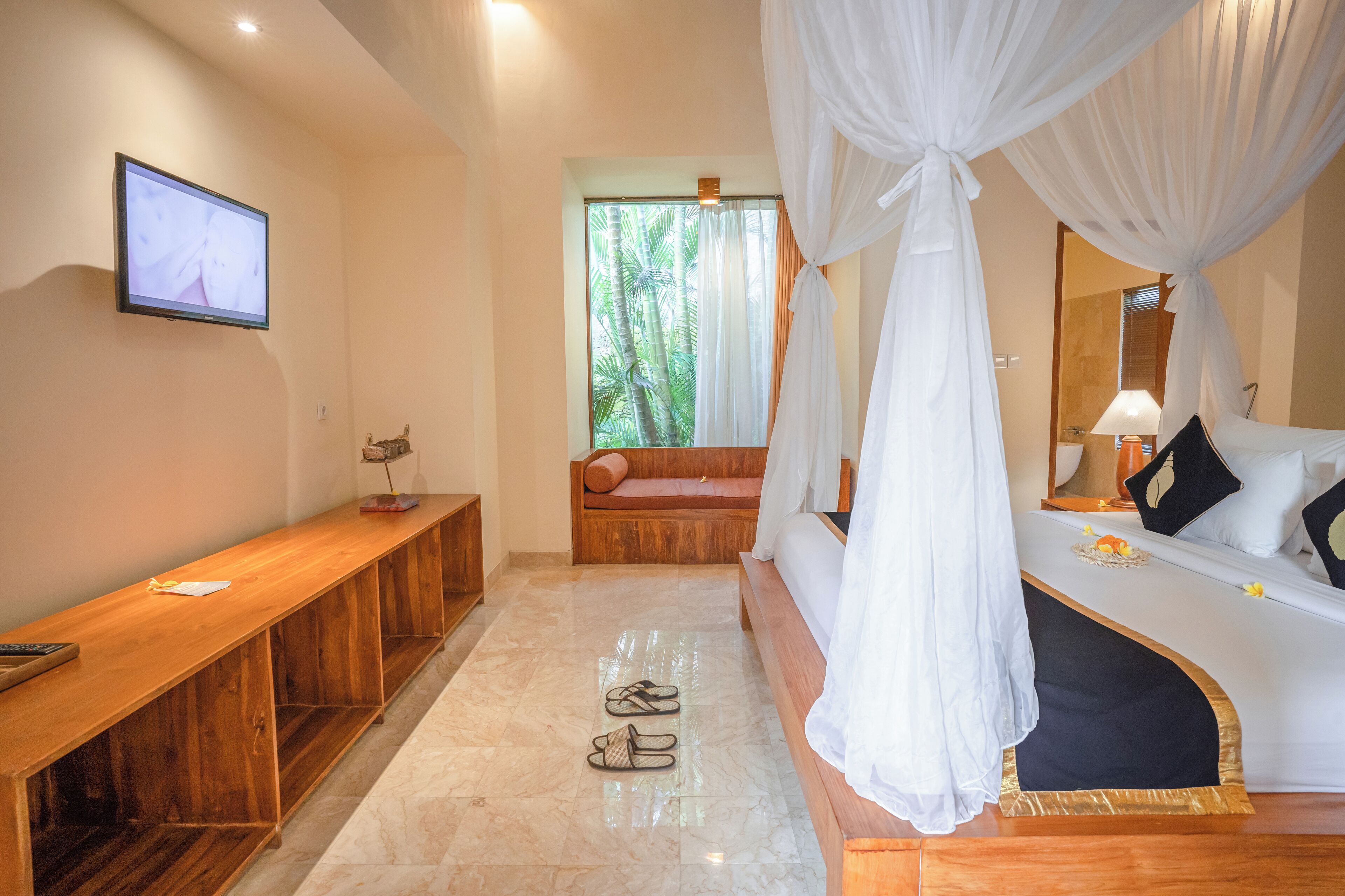 Quarto com um banco de madeira, uma cama com cortinas brancas e uma televisão na parede.