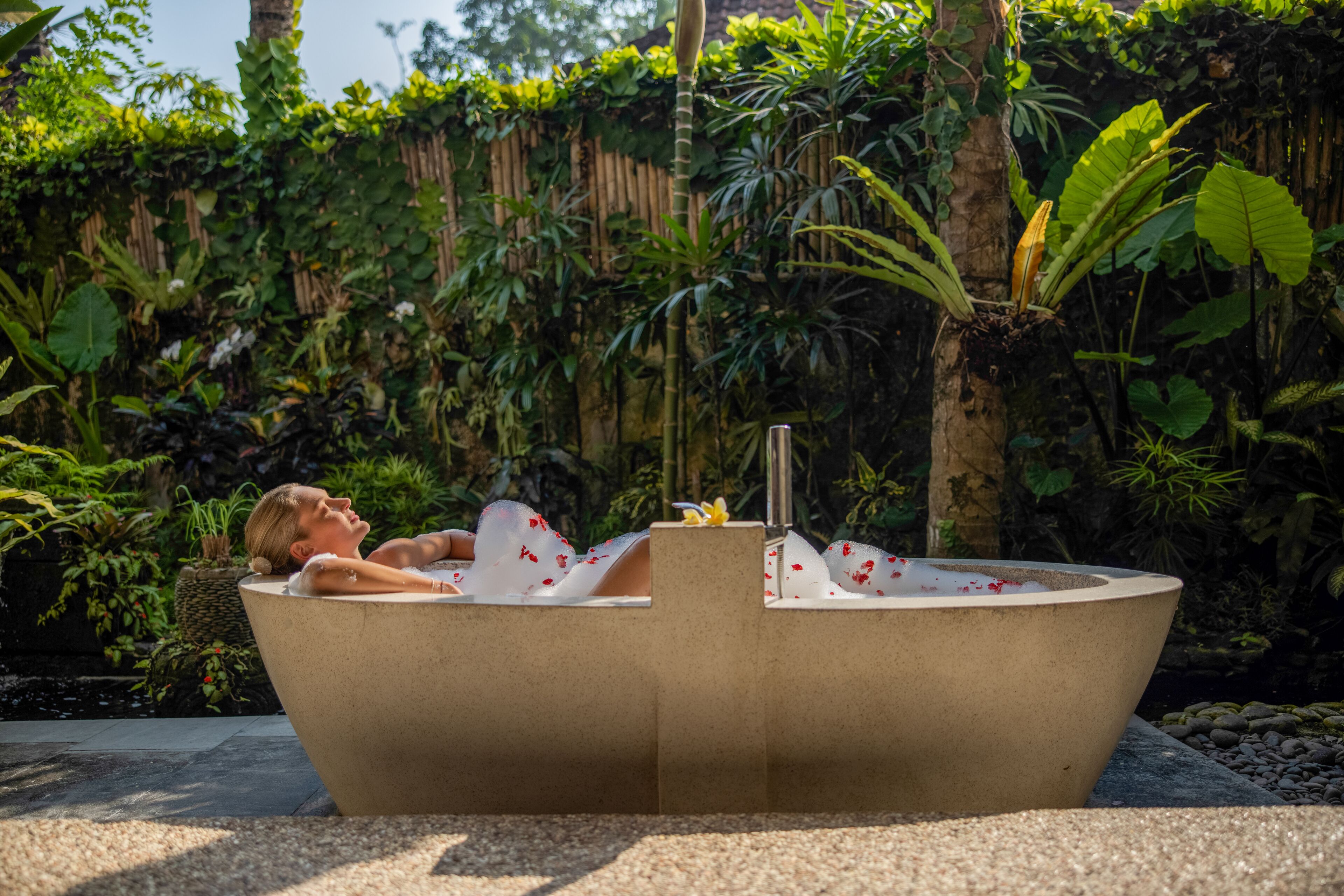 A person relaxing in a bathtub with a plant and a small fountain in a lush garden setting.