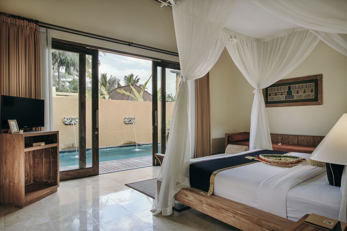 A bedroom with a bed, a TV, a bedside table, a lamp, and a view of a swimming pool.