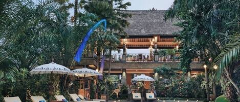 A pool area with lounge chairs and umbrellas, surrounded by lush greenery and a multi-story building with a thatched roof.