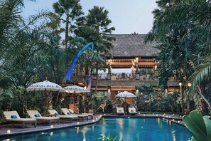 A pool area with lounge chairs and umbrellas, surrounded by lush greenery and a multi-story building with a thatched roof. - The Sankara Resort & Spa (Ubud)