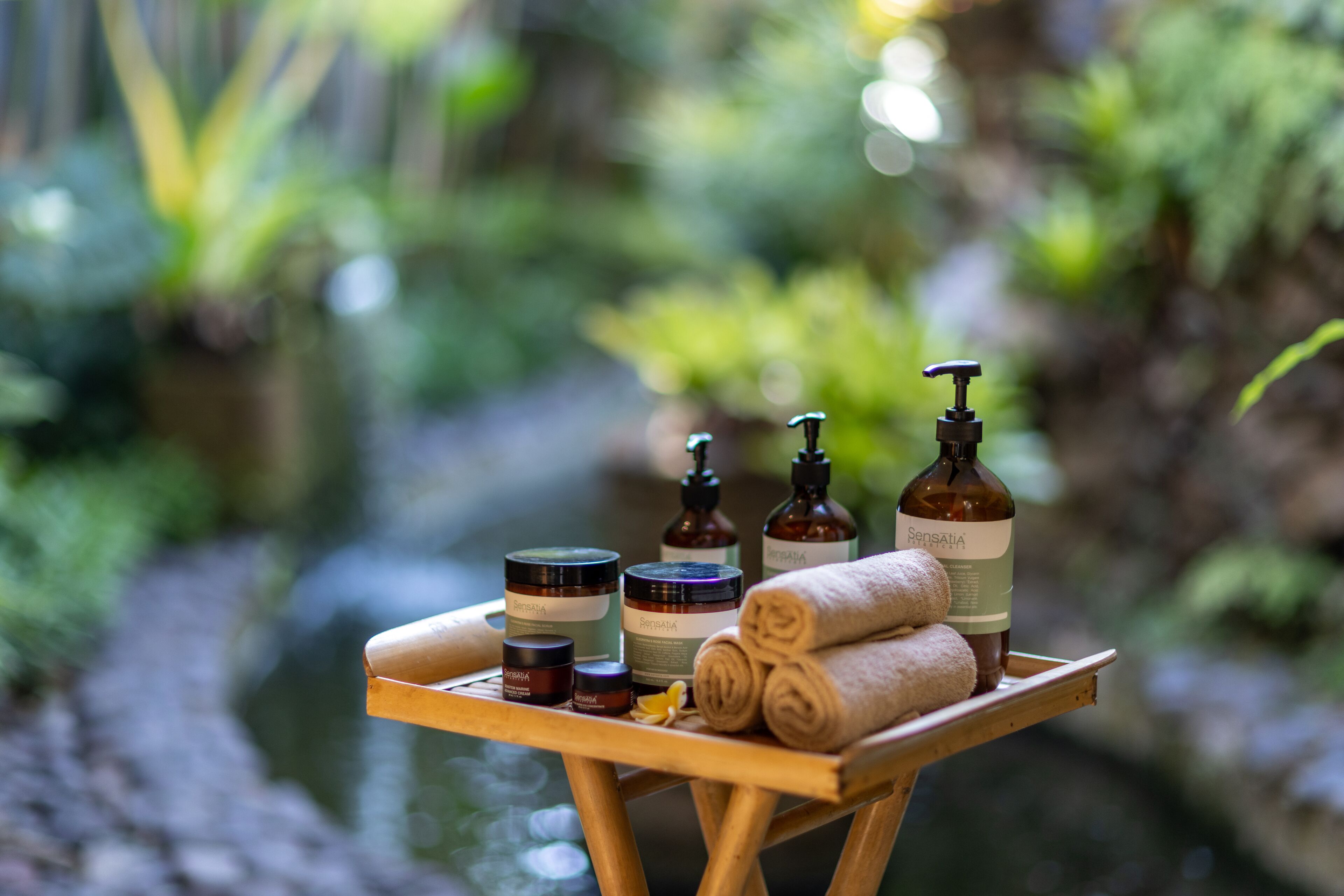 Una bandeja de madera con diversos productos de spa y toallas sobre un soporte de madera en un entorno natural.