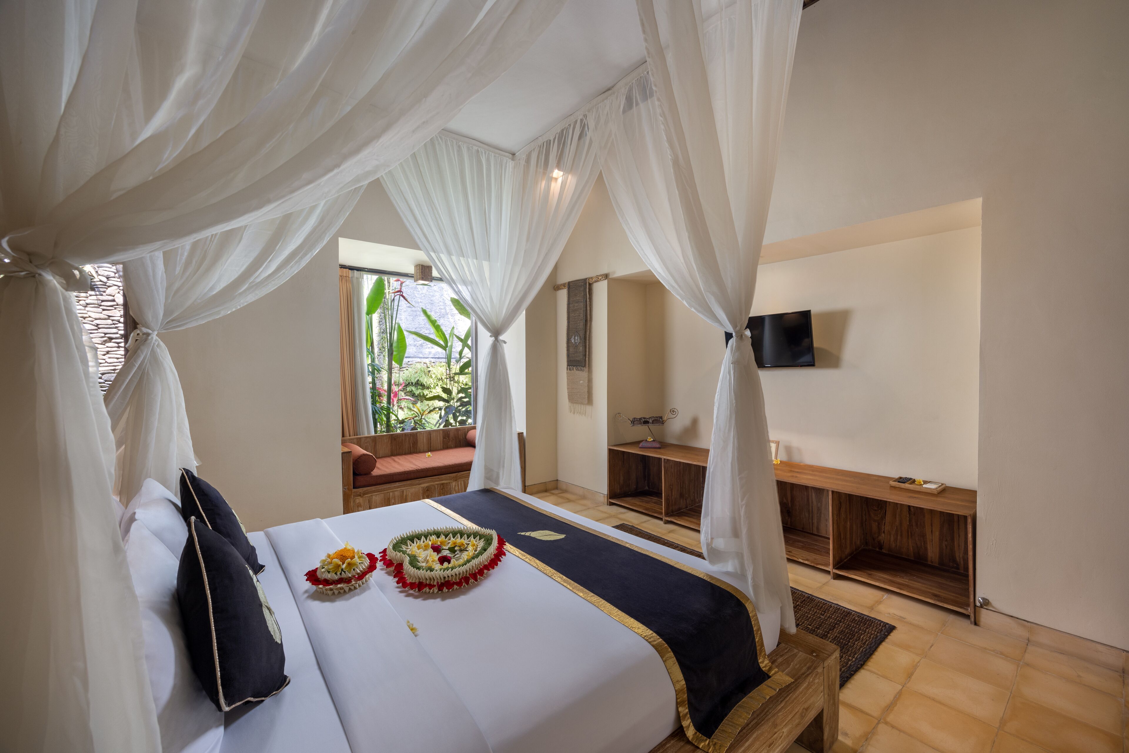 A bedroom with a bed, white curtains, a wooden bench, a TV on the wall, and a window with a view of greenery.