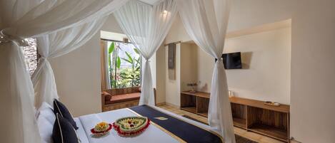 A bedroom with a bed, white curtains, a wooden bench, a TV on the wall, and a window with a view of greenery.