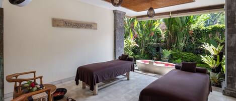 Une salle de spa équipée de deux tables de massage, d’une chaise en bois, d’une petite table agrémentée d’un arrangement floral et d’une grande fenêtre donnant sur un jardin luxuriant.