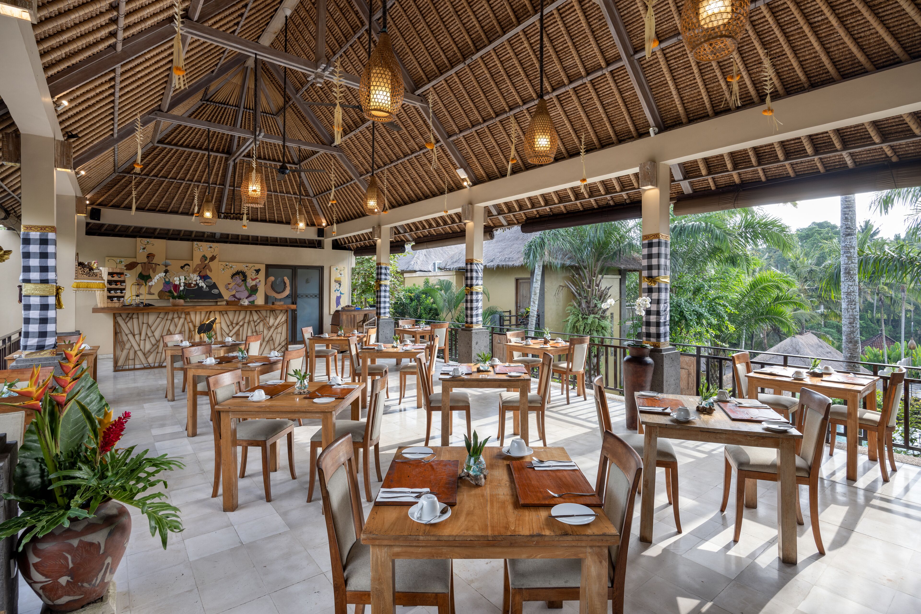A spacious outdoor dining area with wooden tables and chairs, a thatched roof, and potted plants.