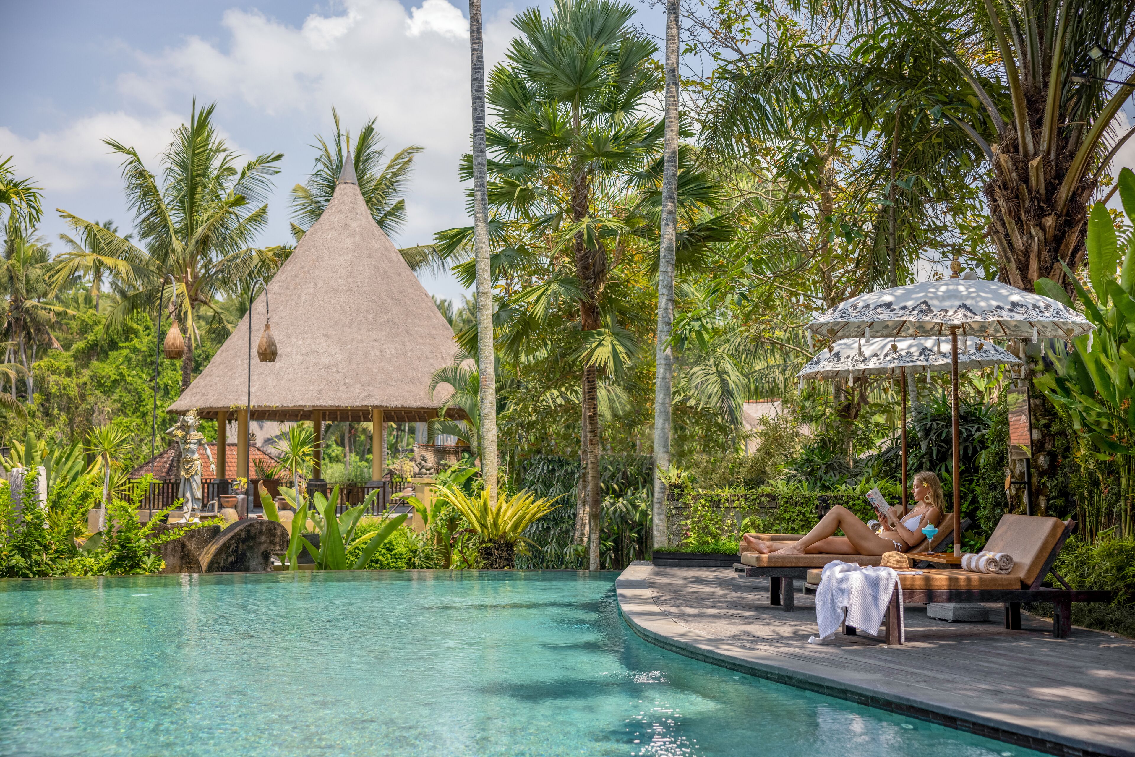 Un área de piscina con sillones de descanso, una estructura de techo de paja y exuberante vegetación tropical.