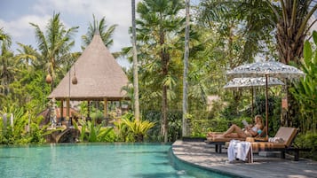 Un área de piscina con sillones de descanso, una estructura de techo de paja y exuberante vegetación tropical.